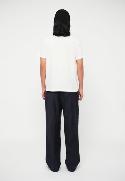 Balta trumpo rankovio marškinėliai derinami su laisvomis tamsiai mėlynomis dryžuotomis kelnėmis, matomos iš nugaros priešais vientisą baltą foną.