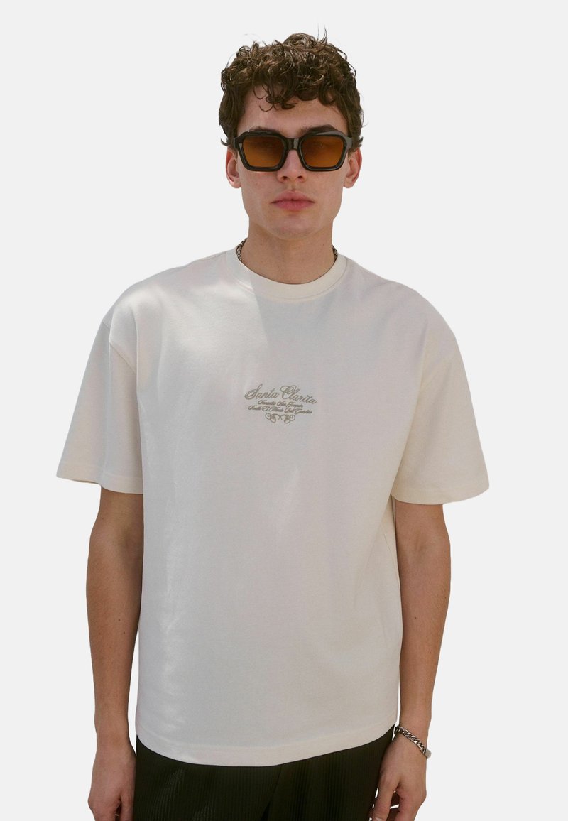 Cremfarbiges T-Shirt mit kurzen Ärmeln und Rundhalsausschnitt, mit dekorativem "Santa Clarita"-Logo in hellbraun, zentral auf der Vorderseite.