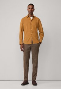 Camisa amarilla de botones con una textura suave, combinada con pantalones marrones de patrón y zapatos oscuros. La camisa tiene un diseño clásico y un cuello.