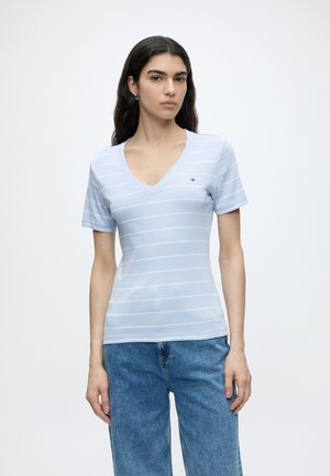 Kvinde med langt mørkt hår iført en lyseblå kortærmet V-hals skjorte med striber og blå jeans mod en ensfarvet hvid baggrund.