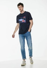 Marinblå t-shirt med grafisk design i röd och blå, kombinerad med ljusblå jeans. Modellen bär vita sneakers och står i en avslappnad pose.