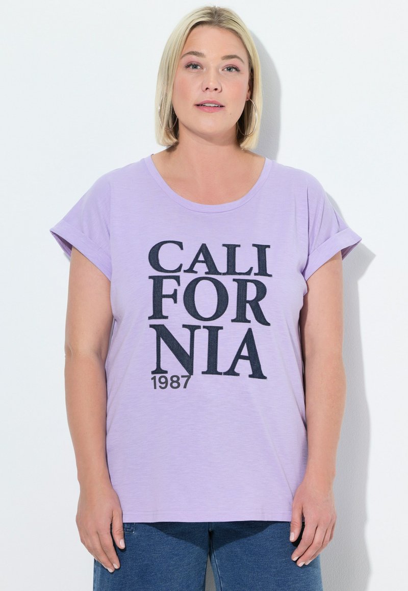 Camiseta de lavanda clara con mangas cortas enrolladas, que presenta un texto negro audaz "CALIFORNIA 1987" en una fuente casual. Tejido suave, corte relajado.