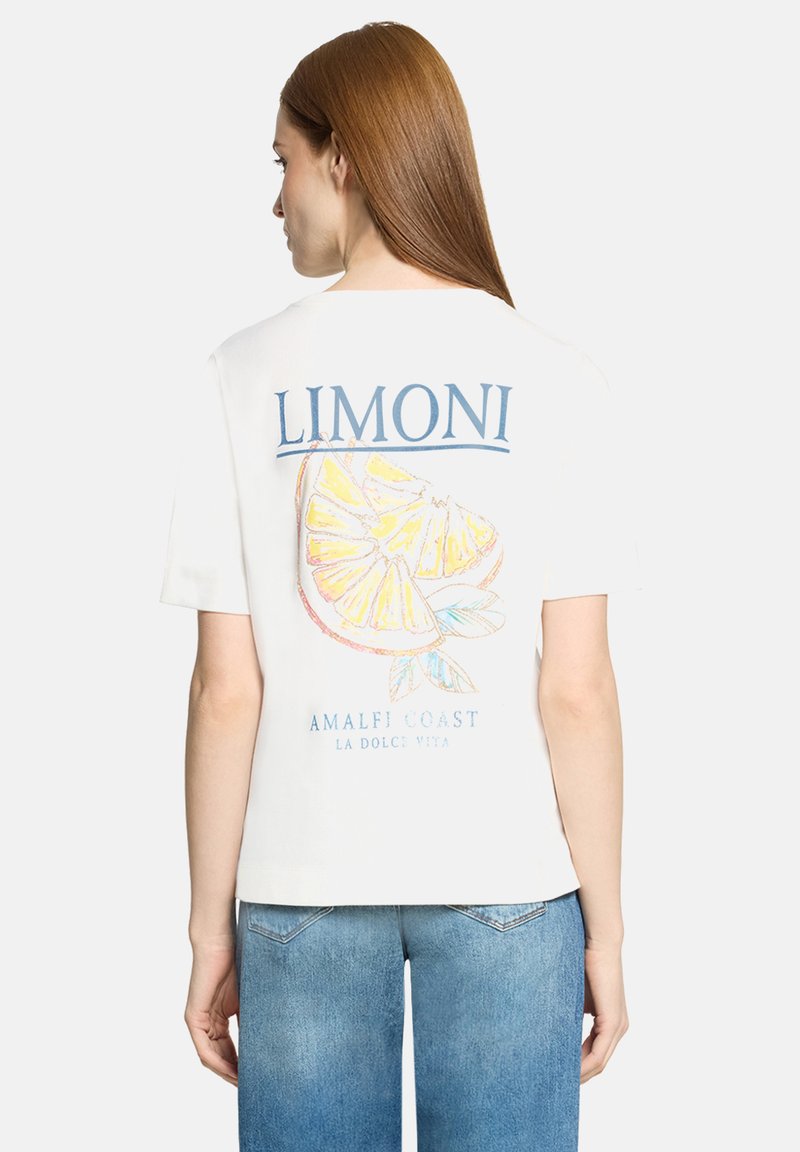Vrouw met lang rood haar die een wit T-shirt draagt met een citroenschijfje als afbeelding en de tekst "LIMONI Amalfi Coast La Dolce Vita" op de achterkant.