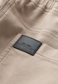 Gros plan sur un tissu beige avec une étiquette noire cousue portant l’inscription « EST. 1985 ombre » sur une couture de poche.