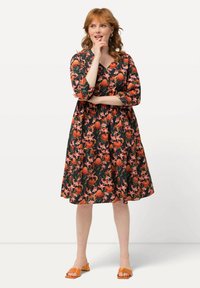 Blumenkleid mit schwarzem Hintergrund. Verfügt über Dreiviertelärmel, V-Ausschnitt und orangefarbene sowie pinke Blumen. Getragen mit orangefarbenen Sandalen.