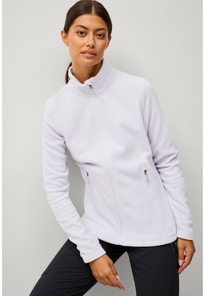 Vrouw met donker haar in een paardenstaart draagt een lichtpaarse fleece jas met ritssluiting en zwarte broek, licht vooroverleunend tegen een effen achtergrond.