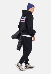 Felpa nera con scritta colorata "ROSSIGNOL", pantaloni della tuta neri, sneakers grigie e un giubbotto imbottito appoggiato sul braccio; berretto di maglia.