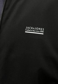 Tissu noir avec texte blanc "JACK & JONES Crafted in blue since 1990" et une fermeture éclair visible sur le côté gauche.