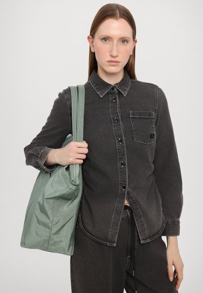 Camisa de ganga preta com fecho de botão, mangas longas e bolso no peito. Segurando uma mala de mão texturizada, verde menta, com duas alças para o ombro.