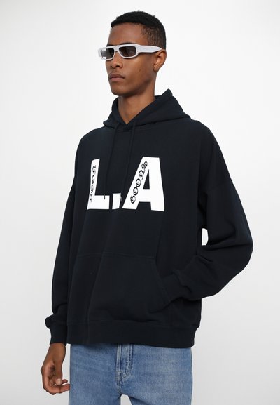 Schwarzer Kapuzenpulli mit großen weißen "L.A."-Schriftzug, Kängurutasche, gerippten Bündchen und lockerer Passform, kombiniert mit hellblauen Jeans.