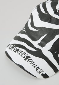 Black and white zebra-striped cap with a curved visor and pronounced "VERSACE JEANS COUTURE" text on the front. Cotton material.
