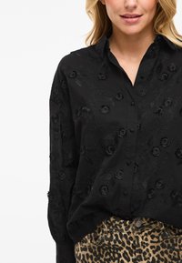 Svart skjorta med knäppning och texturerad broderi av blommor och blad; långärmad, krage och avslappnad passform. Kombinerad med leopardmönstrade byxor.
