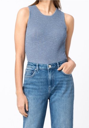 Vrouw die een mouwloze blauwe geribbelde gebreide top draagt, ingestopt in een lichtblauwe, hoog getailleerde spijkerbroek, met één hand in de zak.