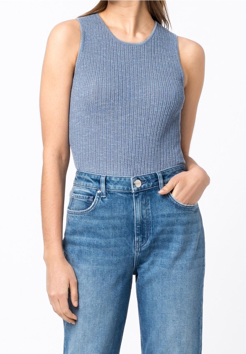 Femme portant un haut sans manches en tricot côtelé bleu, rentré dans un jean denim bleu clair taille haute, une main dans la poche.