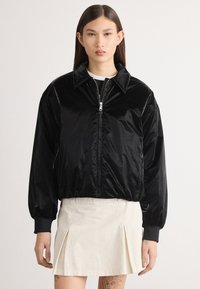 Jachetă bomber neagră, lucioasă, cu fermoar în față, guler mare și manșete elastice. Pur cantă peste un top alb și asociată cu o fustă de culoare deschisă.