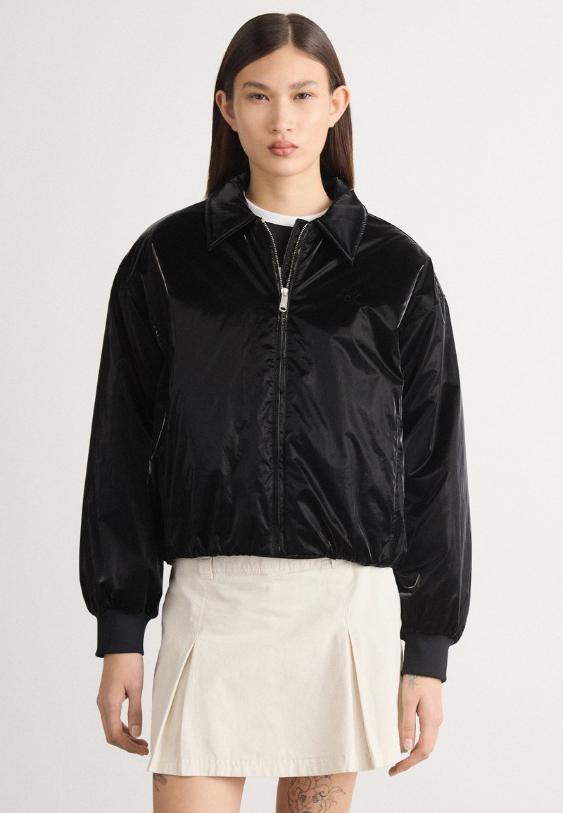 Jachetă bomber neagră, lucioasă, cu fermoar în față, guler mare și manșete elastice. Pur cantă peste un top alb și asociată cu o fustă de culoare deschisă.