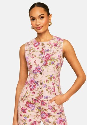 Naine, kes kannab varrukateta roosat lillelist toppi kaardus nööpidega ees ning sobivaid lillelisi pükse, käsi taskus, neutraalne taust.