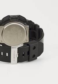 Čierne epoxidové G-SHOCK hodinky so zadnou časťou z nehrdzavejúcej ocele, ktoré majú robustný remienok a nastaviteľné otvory pre lepšie prispôsobenie. Viditeľné strieborné prvky.