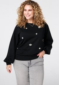 Zwarte sweatshirt met lange, geblazen mouwen en florale versieringen. Zacht materiaal, relaxte pasvorm, geribbelde zoom, gecombineerd met lichtgrijze jeans.