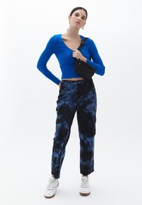 Top a maniche lunghe blu a coste, taglio corto. Pantaloni cargo neri e blu con effetto tie-dye e tasche. Sneakers bianche e una piccola borsa a tracolla nera.