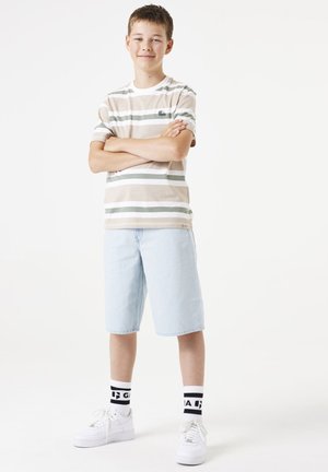 Kurzärmliges gestreiftes T-Shirt in Beige, Weiß und Grün, kombiniert mit hellblauen Jeansshorts und weißen Turnschuhen mit schwarzen Akzenten auf den Socken.