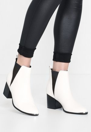 Botas de tobillo de cuero blanco con punta afilada, paneles laterales de elastano negro en contraste y un tacón grueso negro. Accento plateado en la punta.