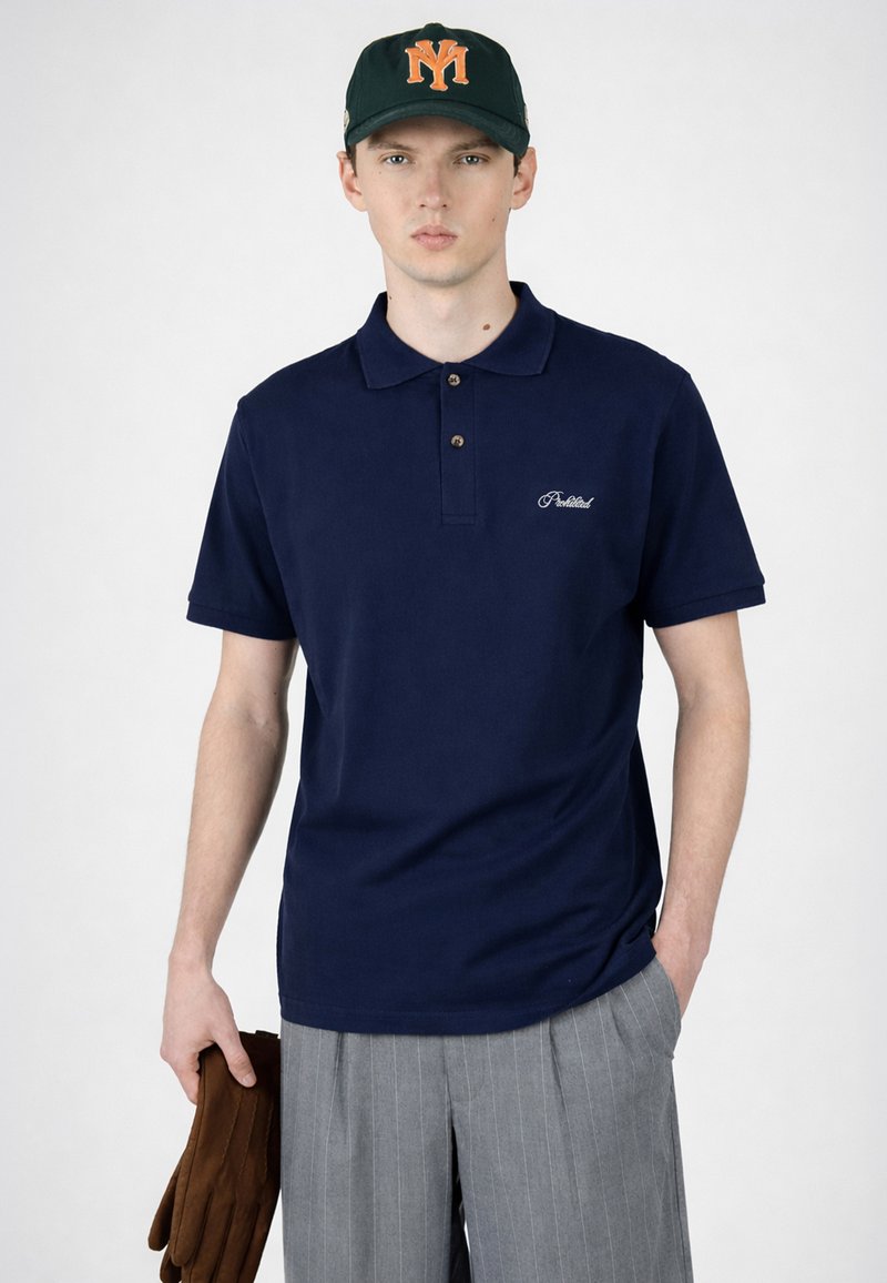 Jeune homme portant un polo bleu marine, un pantalon gris à fines rayures, une casquette vert foncé avec un logo orange, tenant des gants marron dans la main gauche sur fond blanc.