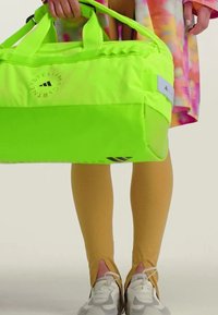 Borsa a tracolla verde neon con manici doppi, caratterizzata da un logo circolare e dettagli neri. Indossata con leggings gialli e sneakers di colore chiaro.