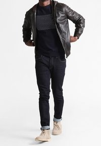 Svart läderbomberjacka över en marinblå tröja med mönstrade detaljer, tillsammans med mörka jeans och beige ankelskor.