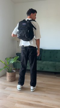 Zwarte stoffen rugzak met een logopatch, gedragen over een wit T-shirt en gecombineerd met zwarte jeans en witte schoenen. Groene plant in de buurt.