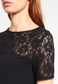 Černé šaty s krátkými krajkovými rukávy se květinovými vzory a jednolitým černým tělem. Krajková textura kontrastuje s hladkým materiálem.