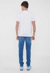 Maglietta bianca a maniche corte, jeans in denim blu e sneakers grigie con accenti blu. Tatuaggio visibile sul braccio sinistro. Design semplice, vestibilità comoda.