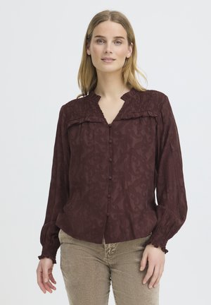 Femme portant un chemisier bordeaux foncé à manches longues avec des boutons sur le devant et un pantalon beige, debout devant un fond clair uni.