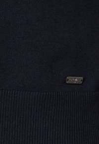 Dunkelblaues Strickpullover mit ribbeltem Saum, weichem Material und einem kleinen metallischen Etikett mit Markennamen am unteren Rand.