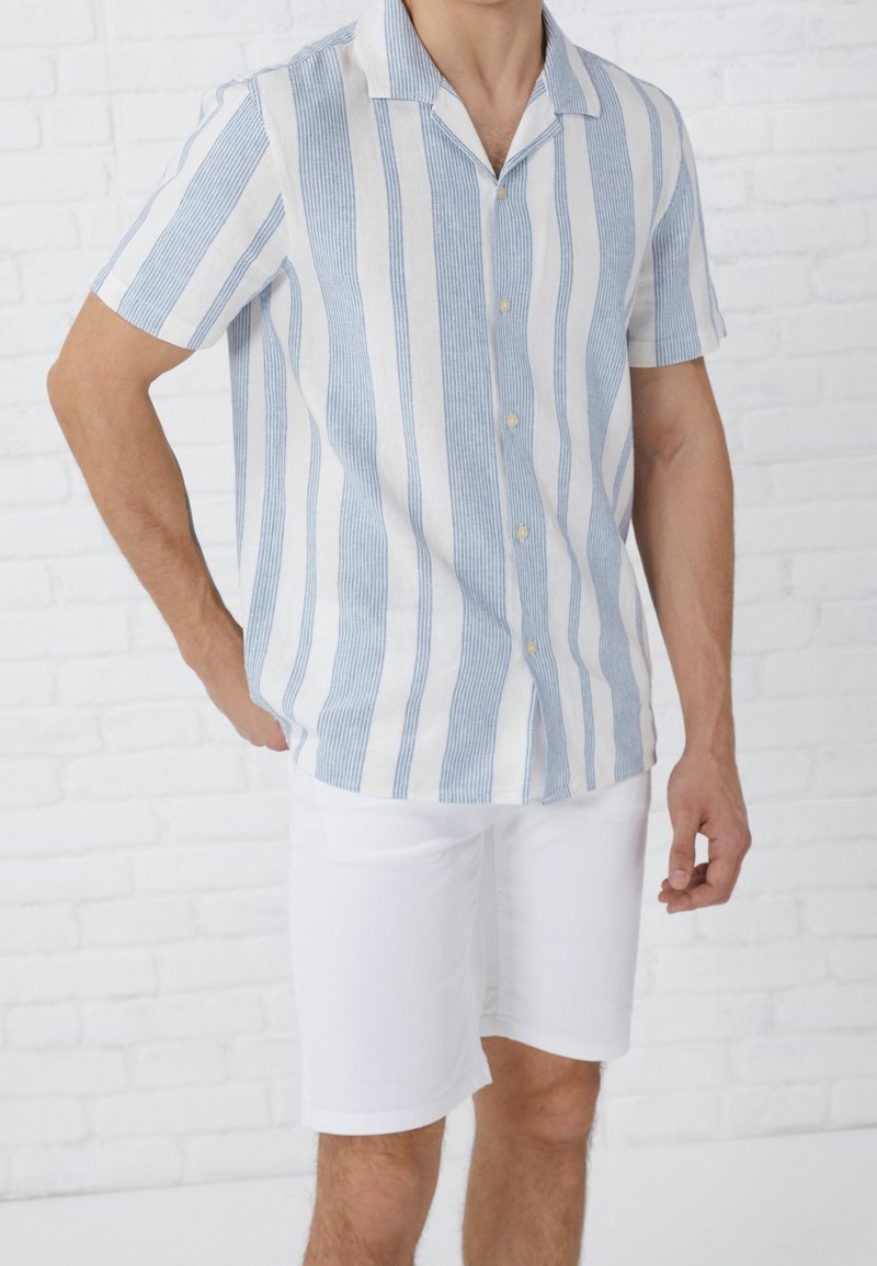 Koszula krótki rękaw w niebiesko-białe pionowe paski, wykonana z lekkiego materiału, z kołnierzem w kształcie notcha i guziczkami, zestawiona z białymi szortami.
