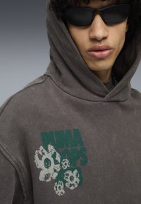 Felpa grigia con un motivo floreale bianco e verde davanti. Presenta un cappuccio con cordino e una vestibilità rilassata. Indossata con occhiali da sole neri.