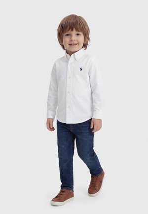 Νέος αγόρι περπατά, φοράει λευκό πουκάμισο με κουμπιά, σκούρα μπλε τζιν και καφέ αθλητικά παπούτσια, σε απλό ανοιχτό φόντο.