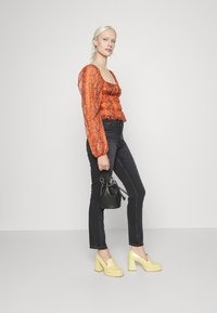 Blusa com padrão de cobra em vermelho e laranja, com mangas bufantes, combinada com jeans justos pretos e saltos grossos amarelos. Modelo segura uma pequena carteira preta.