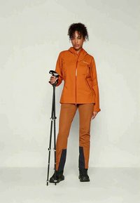 Woman in orange outdoor jacket and pants holding black trekking poles, standing against plain light background.