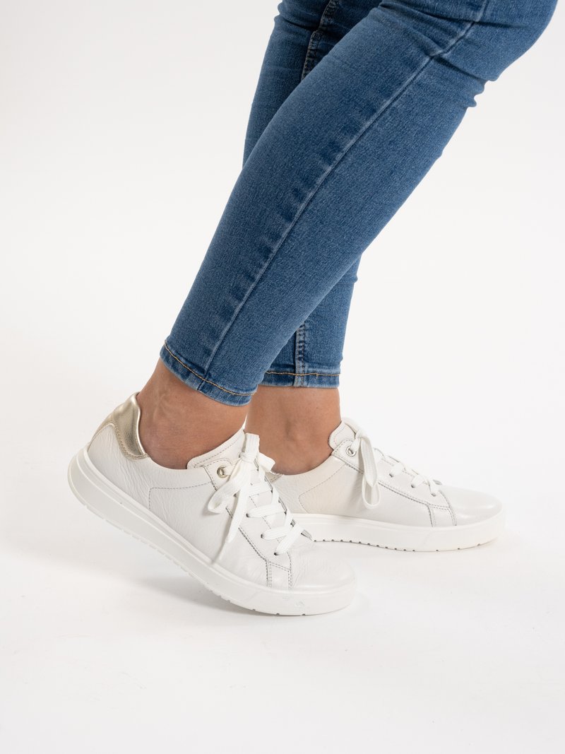 Persona che indossa jeans blu e sneakers bianche con lacci, dettaglio in oro metallizzato sul tallone, in piedi sulle punte dei piedi su sfondo bianco.