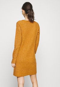 Robe en maille côtelée de couleur moutarde chaude, dotée de longues manches et d'une silhouette décontractée, avec un ourlet légèrement arrondi en bas.