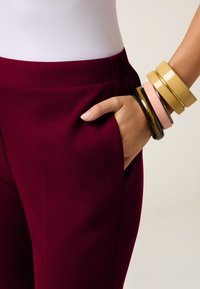 Bordeauxkleurige broek met zijzak, gecombineerd met meerdere armbanden in zwart, geel, roze en schildpadprint. Glad, elastisch materiaal zichtbaar.