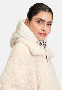 Veste en fausse fourrure de couleur crème avec une texture pelucheuse, un grand col et une capuche blanche rembourrée. Comprend une fermeture éclair avec des accents en métal noir.