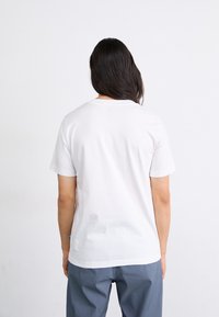 Camiseta blanca de algodón con mangas cortas, cuello redondo y dobladillo recto, exhibida desde la parte de atrás con una textura suave.