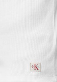 Λευκό ριγωτό ύφασμα με ένα μικρό ορθογώνιο έμπλημα λογότυπου Calvin Klein σε κόκκινο και μαύρο κοντά στο κάτω άκρο.