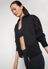 Felpa nera con cappuccio, chiusura a zip davanti, polsini a costine e un piccolo logo sul petto. Abbinata a un reggiseno sportivo nero e leggings.
