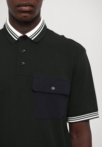 Mørkegrønn polo genser med en brettet krage med hvite og svarte striper, brystlomme og svarte knapper. Teksturert bomullsstoff.