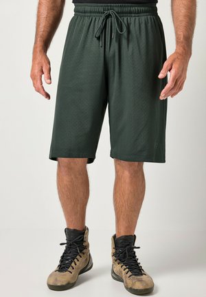 Mand iført mørkegrønne mesh træningsshorts med snøre og højtop-sportsko i beige og sort står på en lys baggrund.