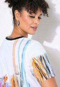 Camiseta blanca con patrones de pinceladas abstractas en colores naranja, azul y negro. Presenta un cuello negro y una textura de tela suave.