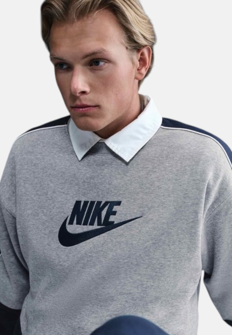 Giovane uomo con capelli biondi che indossa una felpa grigia Nike sopra una camicia bianca con colletto, che guarda leggermente verso il basso e di lato.